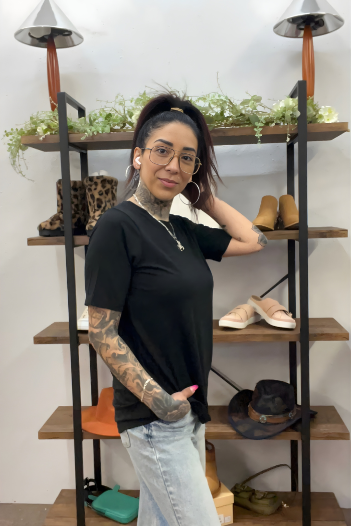 A woman with tattoos, glasses, and a black Basic Short Sleeve Tee stands smiling in front of shelves with shoes, hats, and plants. Her hair is in a high ponytail as she poses with one hand touching her head.