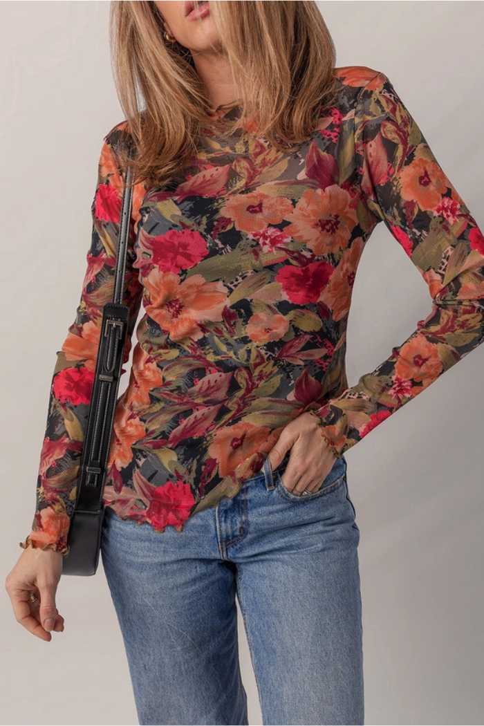 A woman wears the Floral Mesh Lettuce Top, a fitted long sleeve mesh top with red, orange, and green floral patterns. She pairs it with light blue jeans, carries a black shoulder bag, and her light brown hair partially covers her face.