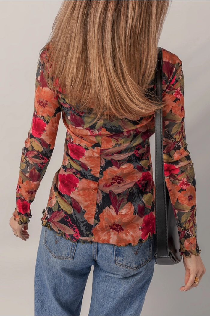 A woman with straight, light brown hair is seen from the back against a plain background, wearing blue jeans and the Floral Mesh Lettuce Top—a long sleeve floral mesh shirt with lettuce trim—paired with a black shoulder bag.