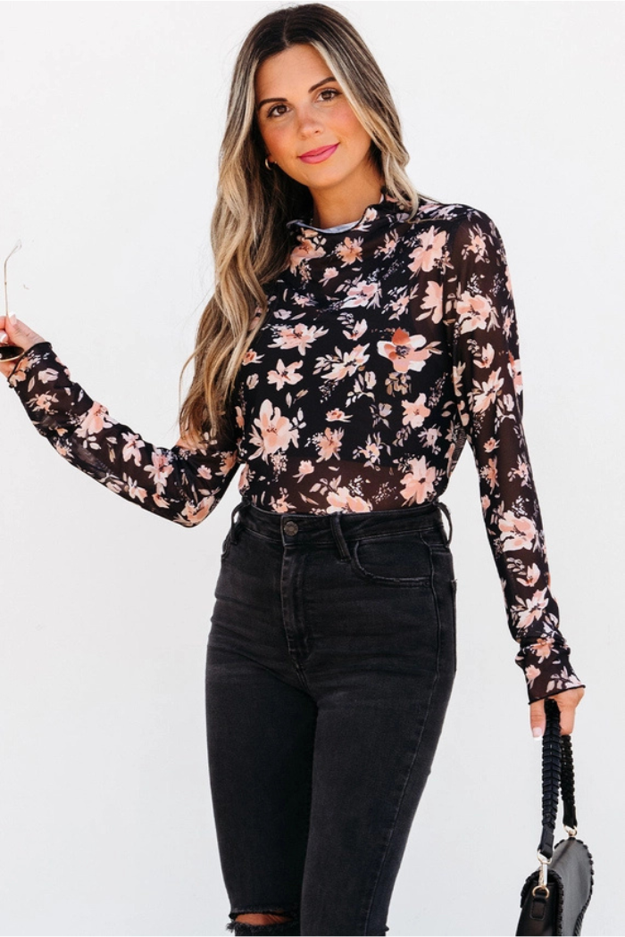 A woman holds a purse, dressed in a chic black Floral Mesh Mock Neck Top with a floral print.