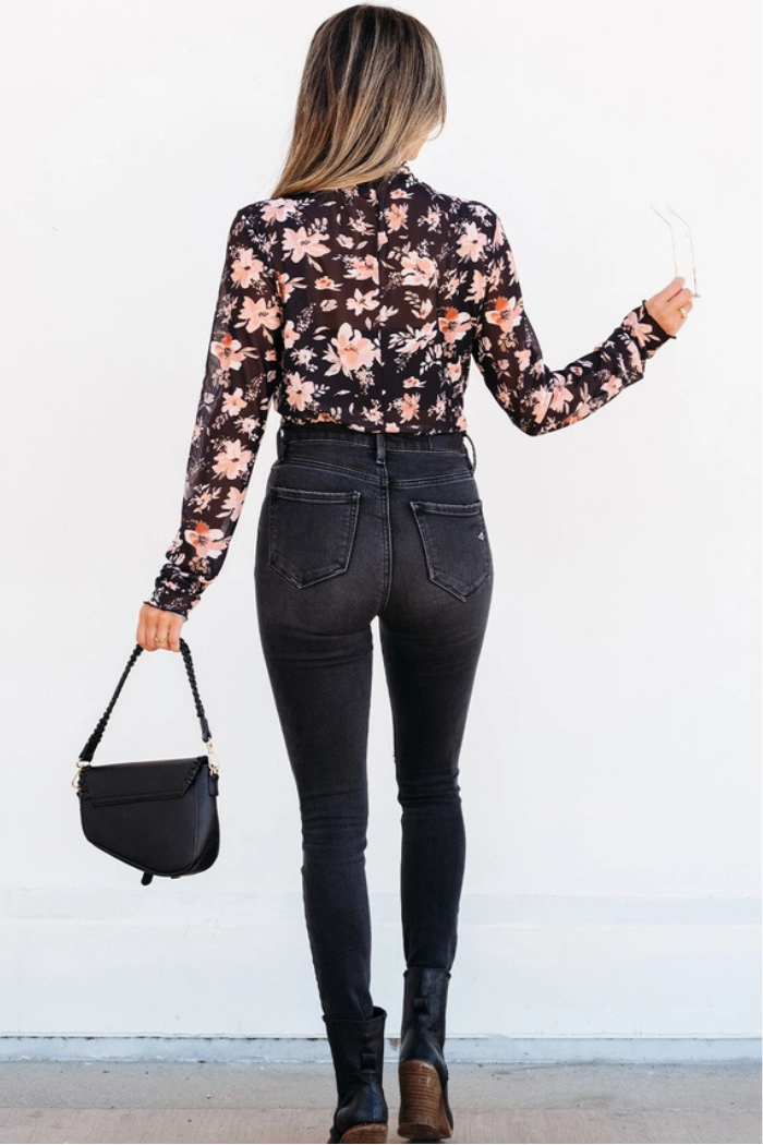 A woman stands facing away, holding a black handbag and sunglasses. She wears the Floral Mesh Mock Neck Top with high-waisted black jeans, black boots, and her long hair cascading down her back.