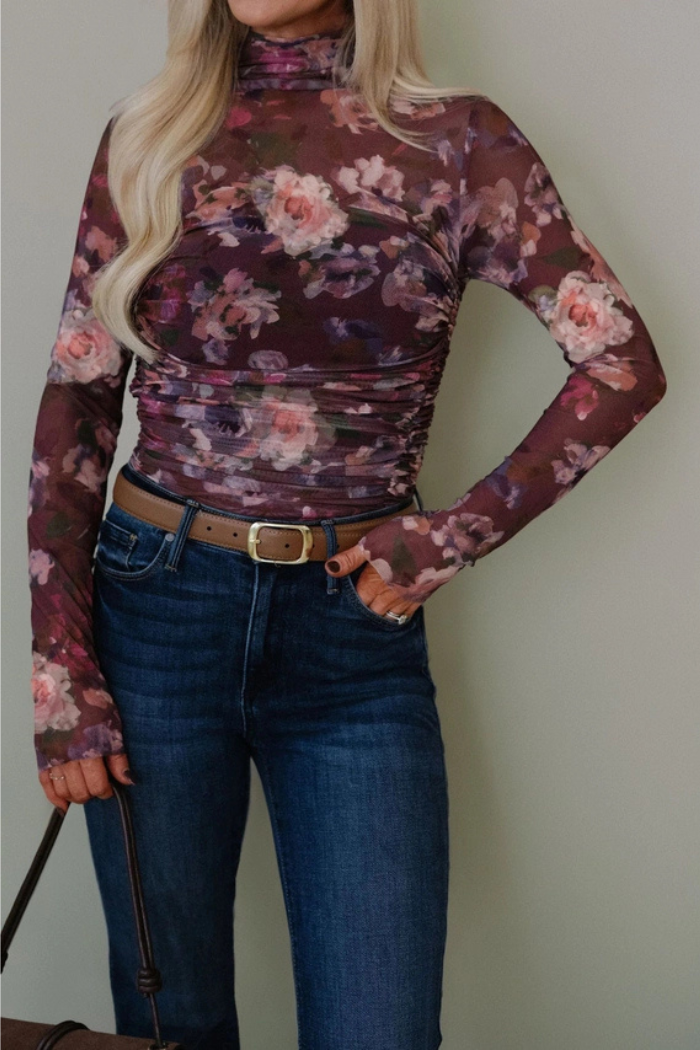 A woman wears a fitted, long-sleeve Floral Ruched Turtleneck Top in purple and pink mesh, paired with high-waisted blue jeans, a brown belt, and a bag. Her blonde hair is styled in loose waves.