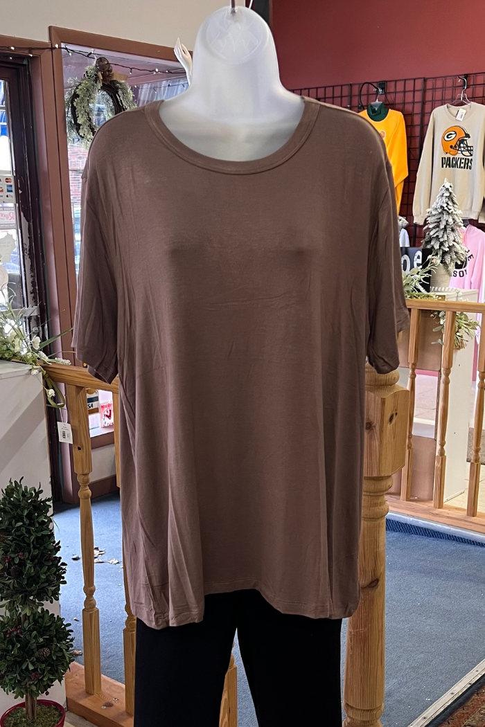A Plus Size Rayon Tee in brown is shown on a white mannequin torso inside a store, with wooden railings and sports shirts hanging in the background.