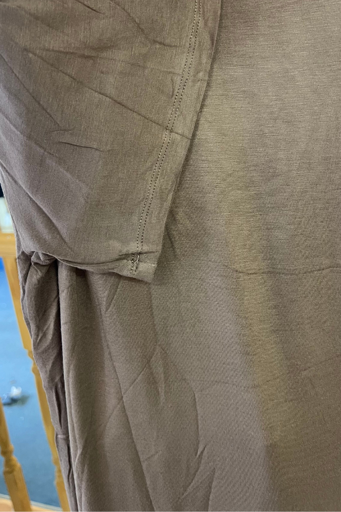 A close-up of a wrinkled, light brown Plus Size Rayon Tee highlights the sleeve and side seam, with part of a staircase railing and carpeted floor in the background.