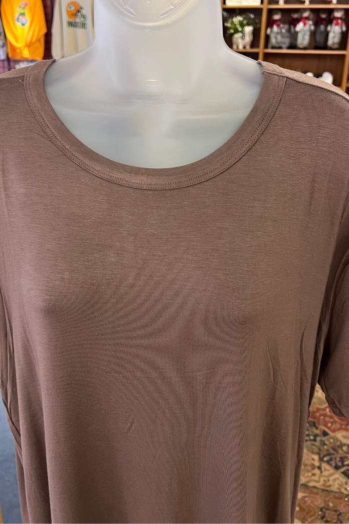 A Plus Size Rayon Tee from Zenana Premium, brown with short sleeves and made from a soft rayon blend, is shown on a mannequin torso. Shirts and shelves with various items appear in the background.
