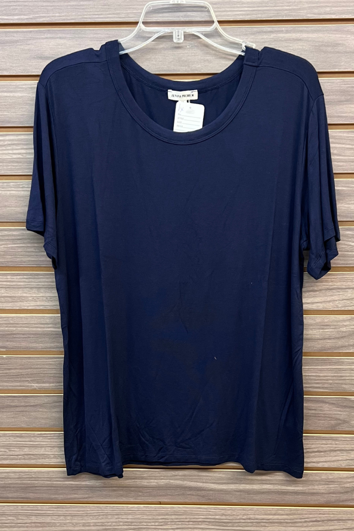 A Plus Size Rayon Tee in plain navy blue from Zenana Premium hangs on a clear plastic hanger against a wooden slat wall. A white tag at the neckline highlights the soft rayon blend comfort of this short-sleeve tee.