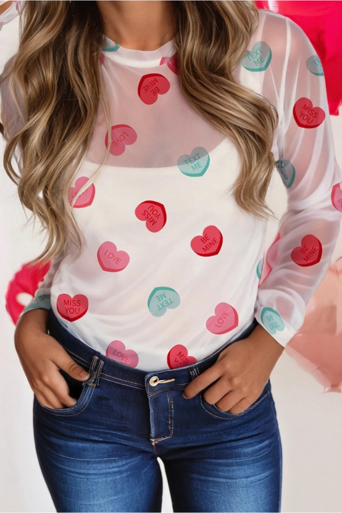 A woman wears the Heart Mesh Valentine Top, a sheer blouse with colorful candy heart prints saying LOVE, MISS YOU, and BE MINE. Paired with blue jeans, her hand rests in her pocket for a playful look.