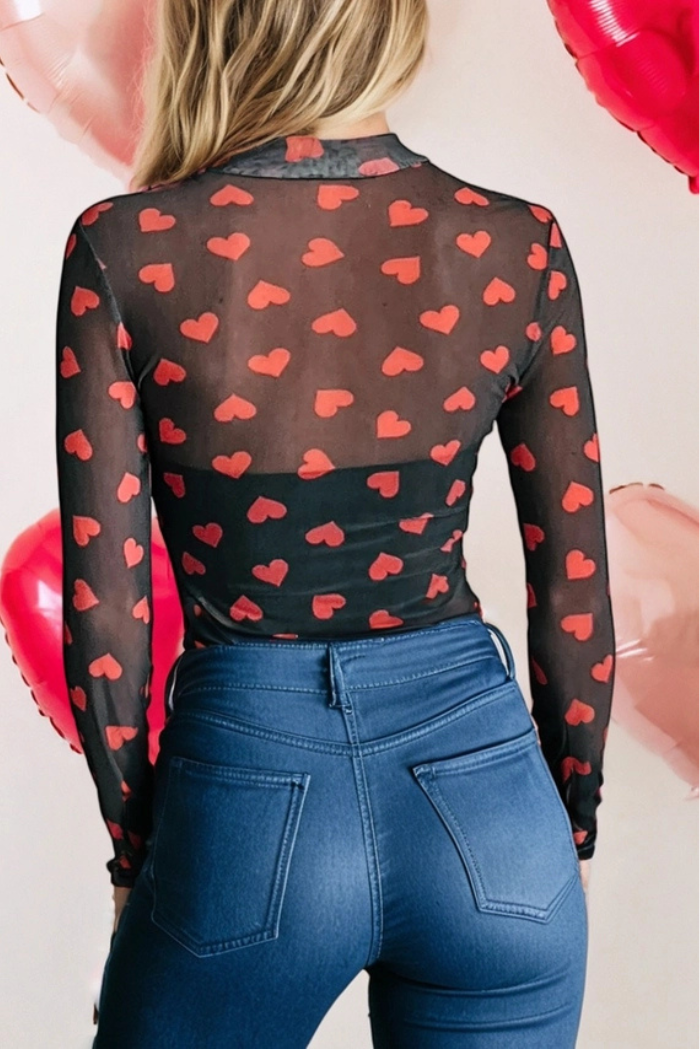 A woman shown from the back wears the Valentine Heart Mesh Top with a red heart pattern and high-waisted blue jeans. Pink and red heart-shaped balloons are in the background.