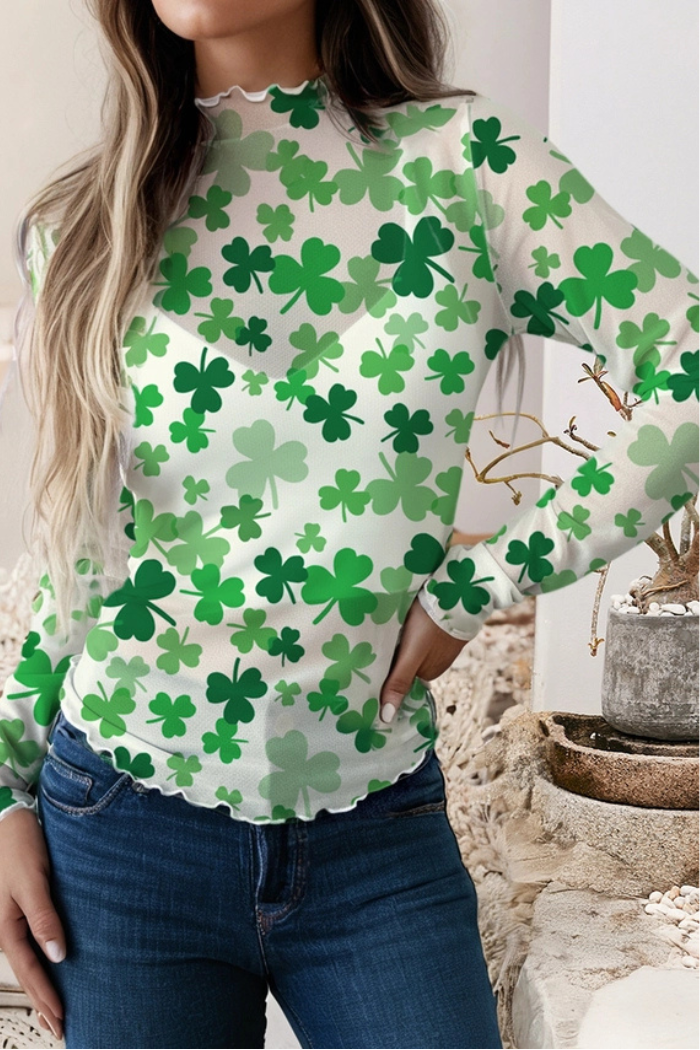 A woman confidently models the St. Patrick Mesh Top, featuring mesh fabric with green shamrock patterns. She pairs this festive top with blue jeans, her long hair cascading over her shoulders as she stands with a hand on her hip.