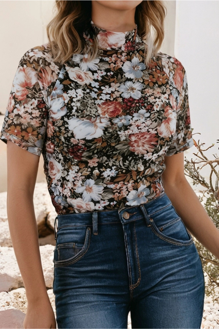Outdoors, a person wears high-waisted blue jeans with a Floral High Neck Mesh Top featuring short sleeves and a mix of white, brown, and pink flowers. Their face is not visible.
