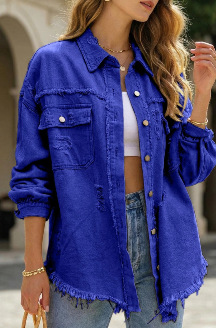 Wearing the Ripped Edge Denim Jacket in bright blue over a white crop top and light blue jeans, accessorized with gold jewelry, a woman stands outdoors.