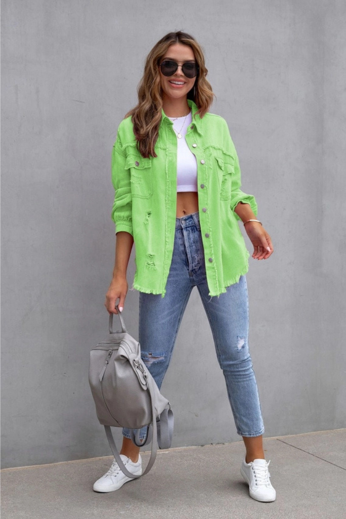 A woman smiles at the camera, wearing a Ripped Edge Denim Jacket over a white crop top, blue ripped jeans, and white sneakers. She holds a gray backpack, has wavy hair, and wears sunglasses while standing against a gray wall.