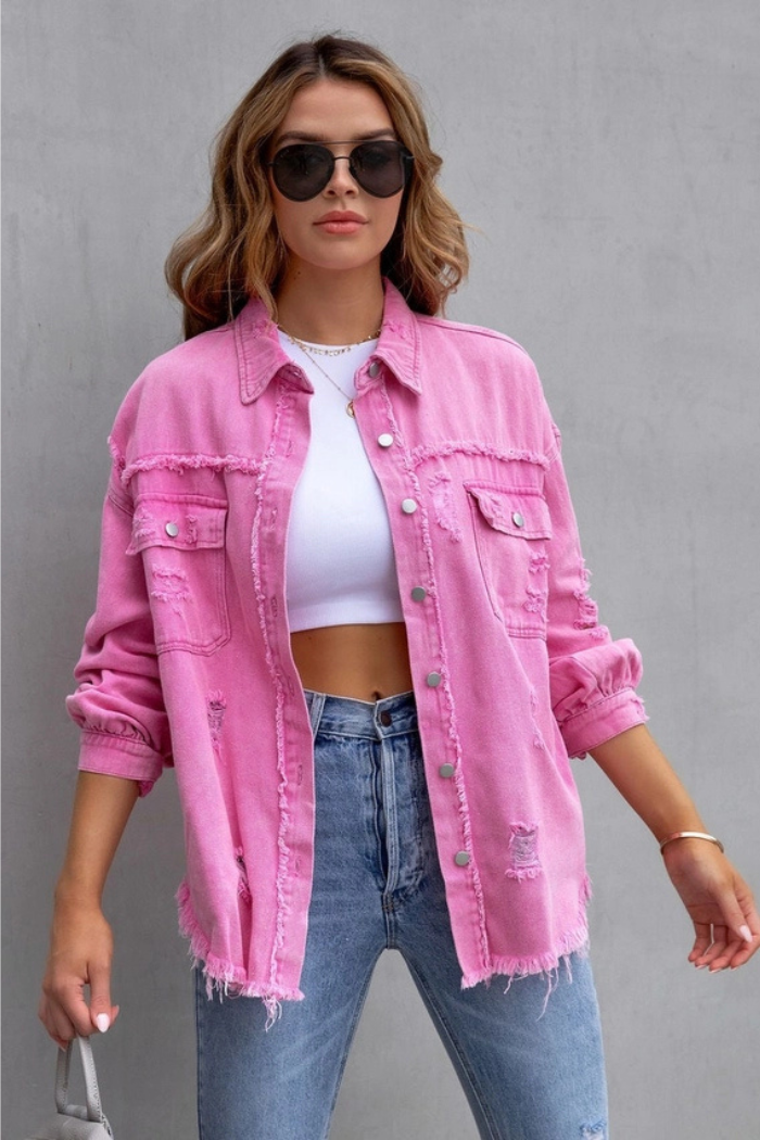 A woman models the Ripped Edge Denim Jacket in bright pink over a white crop top with light blue jeans, wavy hair, and large black sunglasses, standing against a plain gray background.