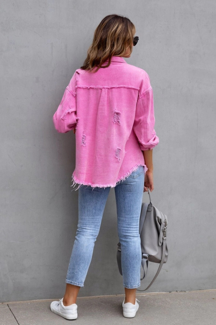A woman faces a gray wall, wearing the Ripped Edge Denim Jacket in pink, light blue cropped jeans, and white sneakers. She holds a light gray backpack in her left hand and has loose hair.