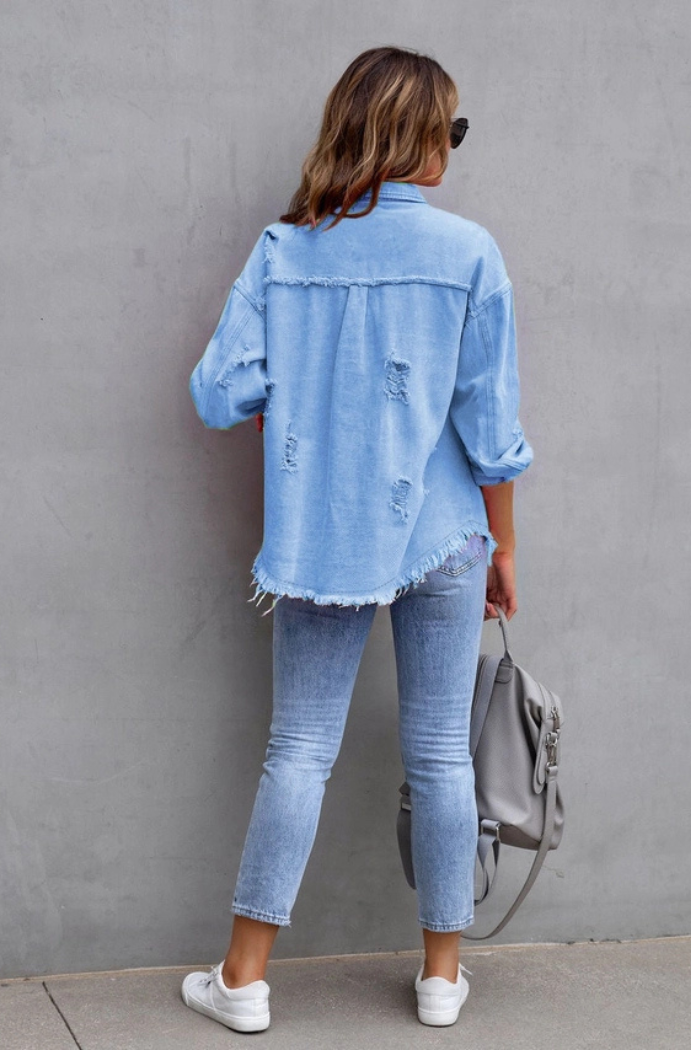 A person with wavy hair faces a gray wall, wearing a Ripped Edge Denim Jacket, matching jeans, white sneakers, and holding a gray bag.