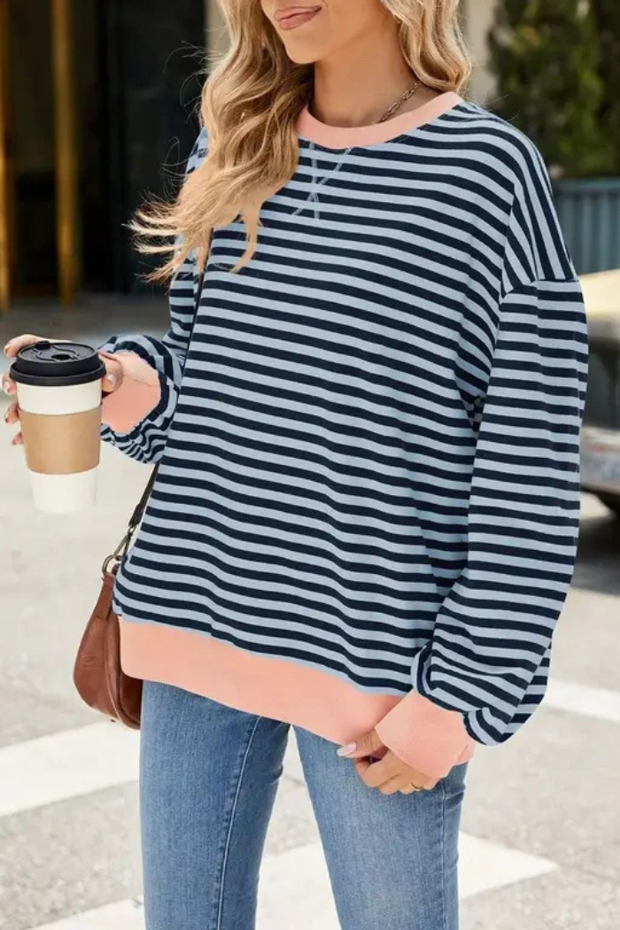 A woman, face not visible, stands outdoors holding a takeaway coffee and wearing blue jeans with the Relaxed Stripe Sweatshirt featuring pink cuffs and hem.