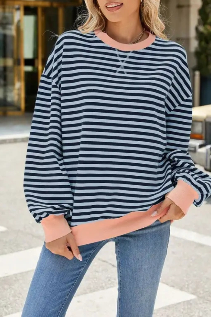 Outdoors, a woman wears the Relaxed Stripe Sweatshirt—navy blue and white stripes with light pink cuffs and collar—paired with blue jeans.