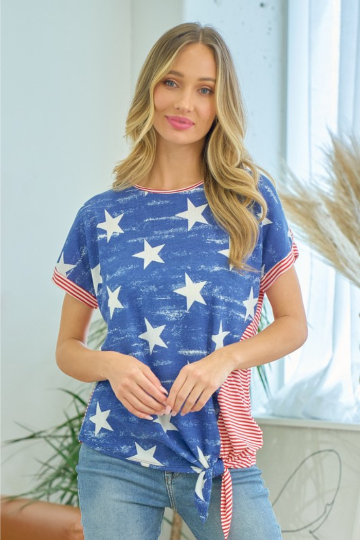 A woman with long blonde hair wears the Stars & Stripes Top—a casual knit with white stars, red and white striped sleeves—paired with blue jeans, standing indoors near a window and some plants.