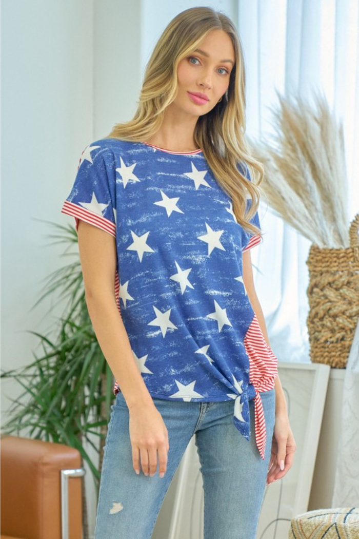 A woman with long blonde hair wears the Stars & Stripes Top, featuring white stars, red and white striped trim, and a side tie, paired with light blue jeans. She stands indoors in a bright room with plants and neutral decor.