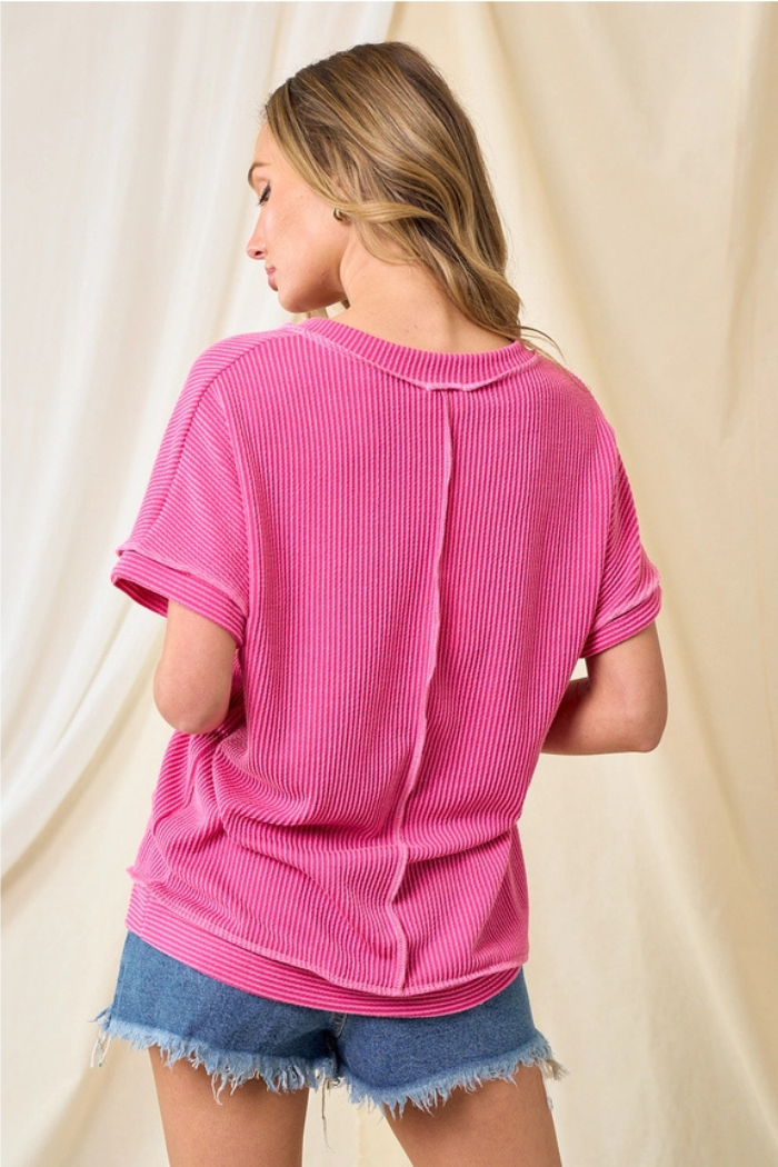 A woman with long, wavy blonde hair wears an oversized Basic Ribbed V-Neck Top in pink and frayed denim shorts, standing with her back to the camera against a cream fabric backdrop—ideal for everyday style.