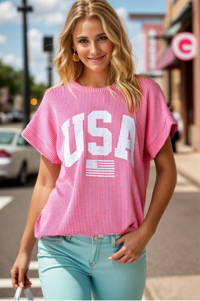 A smiling woman stands outside on a city street wearing the USA Ribbed Dolman Top, featuring a pink ribbed design with a small white American flag, paired with light blue jeans for a casual patriotic vibe.