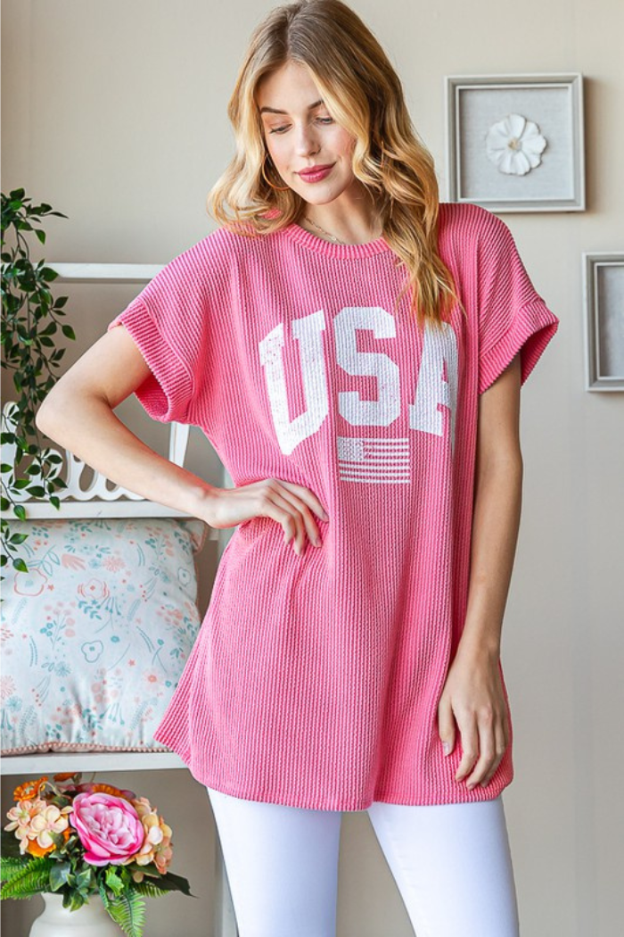 A woman with long blonde hair wears the USA Ribbed Dolman Top in pink with white lettering, paired with white leggings. She stands indoors by a floral cushion and small plant, smiling and looking down.