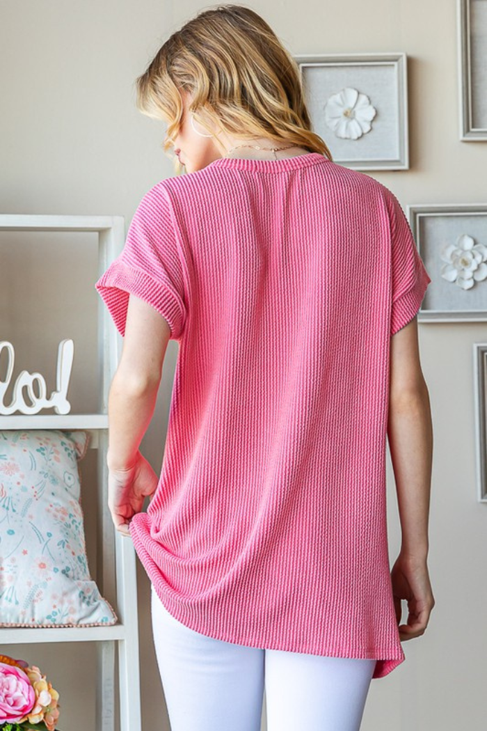A woman with blonde hair wears the USA Ribbed Dolman Top styled as a loose pink shirt with white pants, standing indoors facing away from the camera in a room decorated with floral art and soft furnishings.