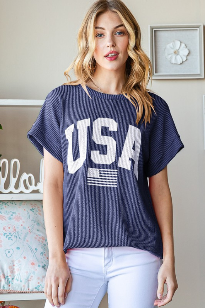 A blonde woman wears the Plus Size USA Top in navy blue with white letters. She stands indoors by a beige wall with floral decor. The waffle-textured knit of the shirt offers extra style and comfort.