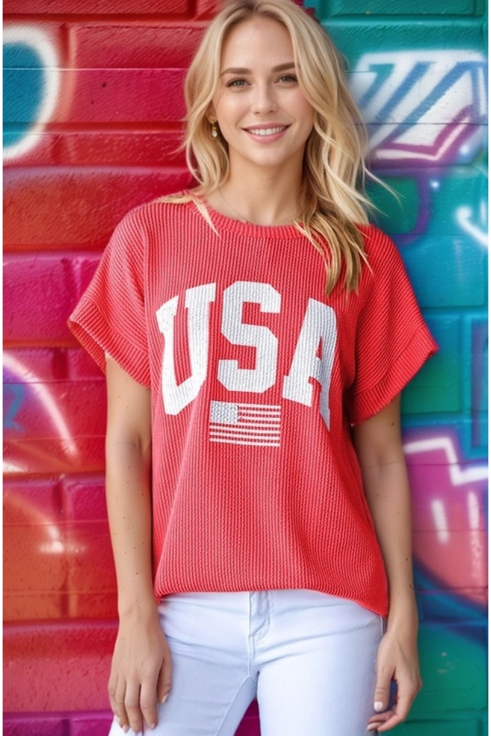 A smiling woman with long blonde hair wears the Plus Size USA Top and white pants while standing before a vibrant graffiti wall.