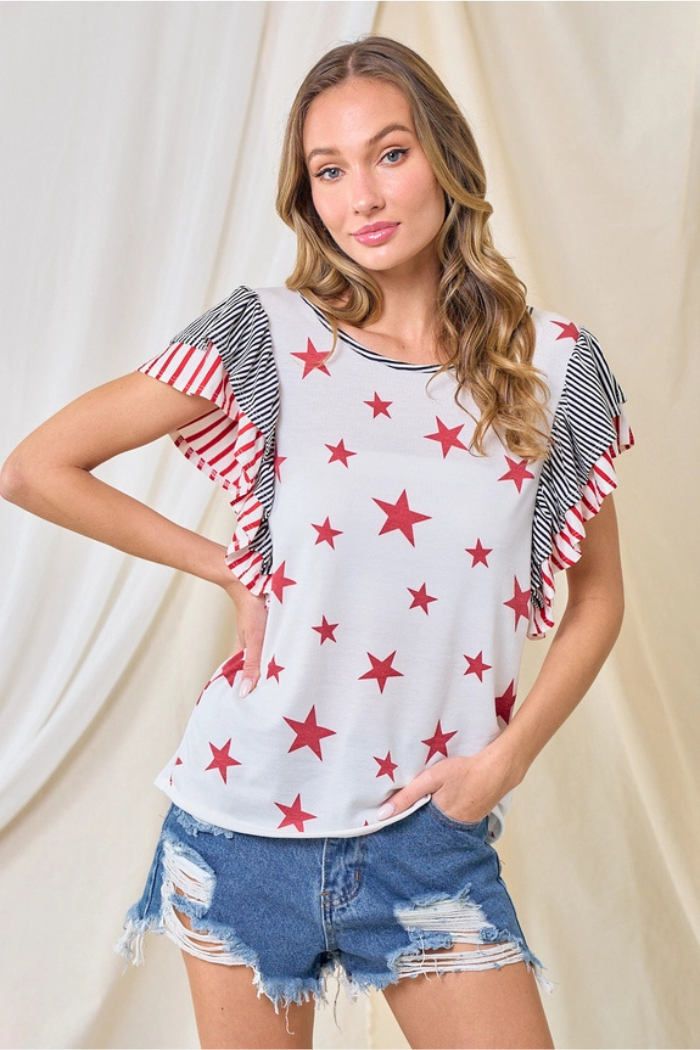 A woman wearing the Party in the USA Top with red stars, ruffled striped sleeves, and distressed denim shorts poses against a neutral draped fabric backdrop with one hand on her hip.