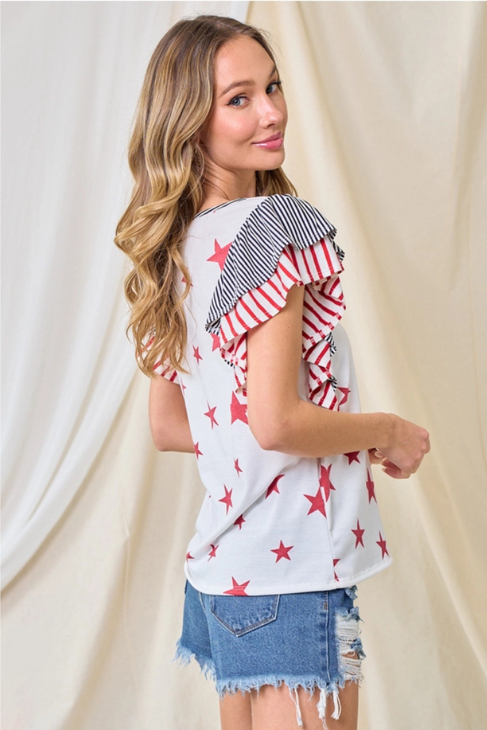 A woman with long wavy hair smiles as she turns to the side, wearing frayed denim shorts and the Party in the USA Top featuring patriotic ruffle sleeves with red stars, black and white stripes, and red and white stripes.