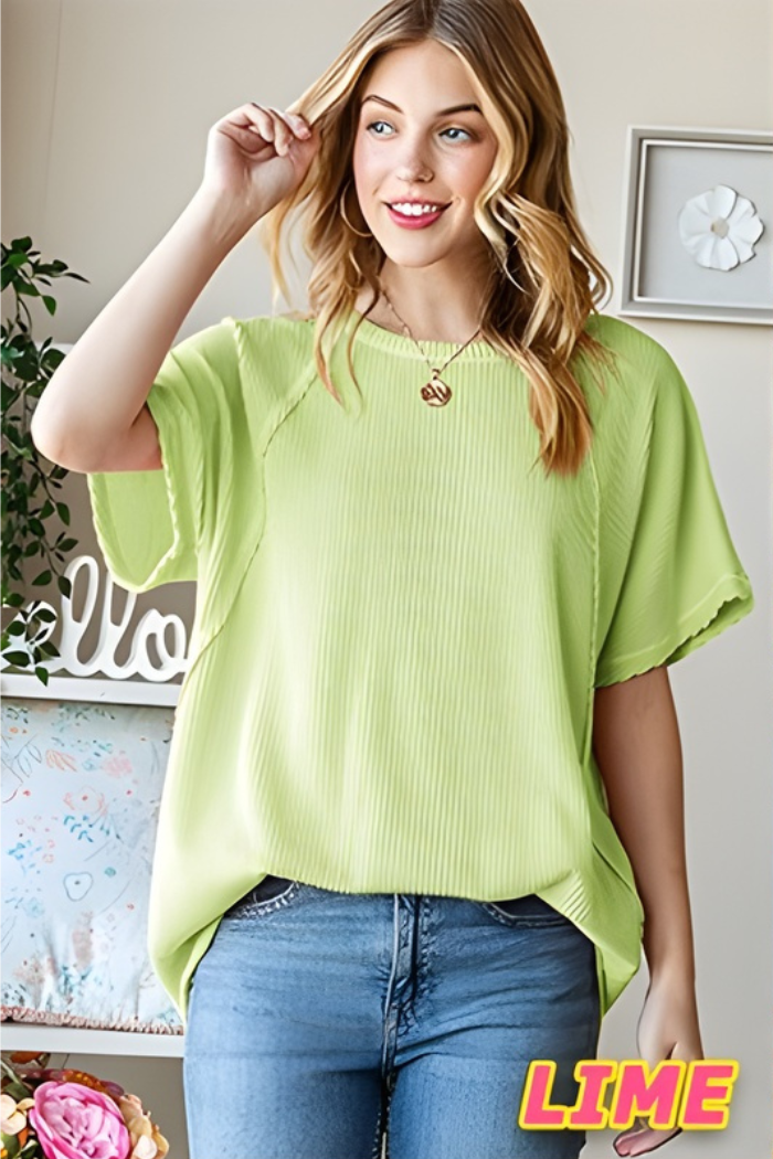 A smiling blonde woman wears the Solid Urban Ribbed Top in loose, light lime green with an oversized fit and blue jeans. She stands indoors near a pillow and plants. "LIME" appears in bold yellow and pink text in the corner.