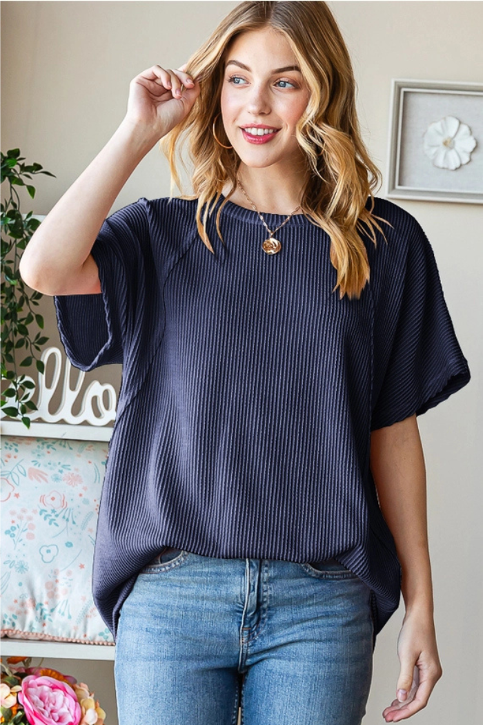 A smiling woman with wavy blonde hair stands indoors with one hand near her head, wearing a Solid Urban Ribbed Top and blue jeans. A floral pillow and plant are visible in the background.