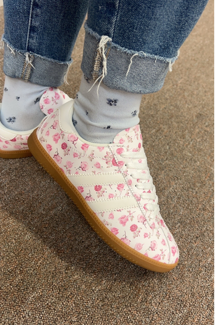 Someone wearing frayed cuffed jeans and white socks with blue accents stands on a brown carpet, showcasing the Floral Lace-Up Sneakers—women’s shoes featuring pink floral patterns and gum soles for a stylish casual look.