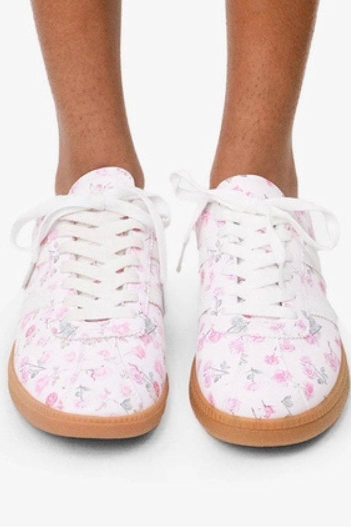 A person stands on a white background wearing the Floral Lace-Up Sneakers, which feature pink floral patterns and white laces.