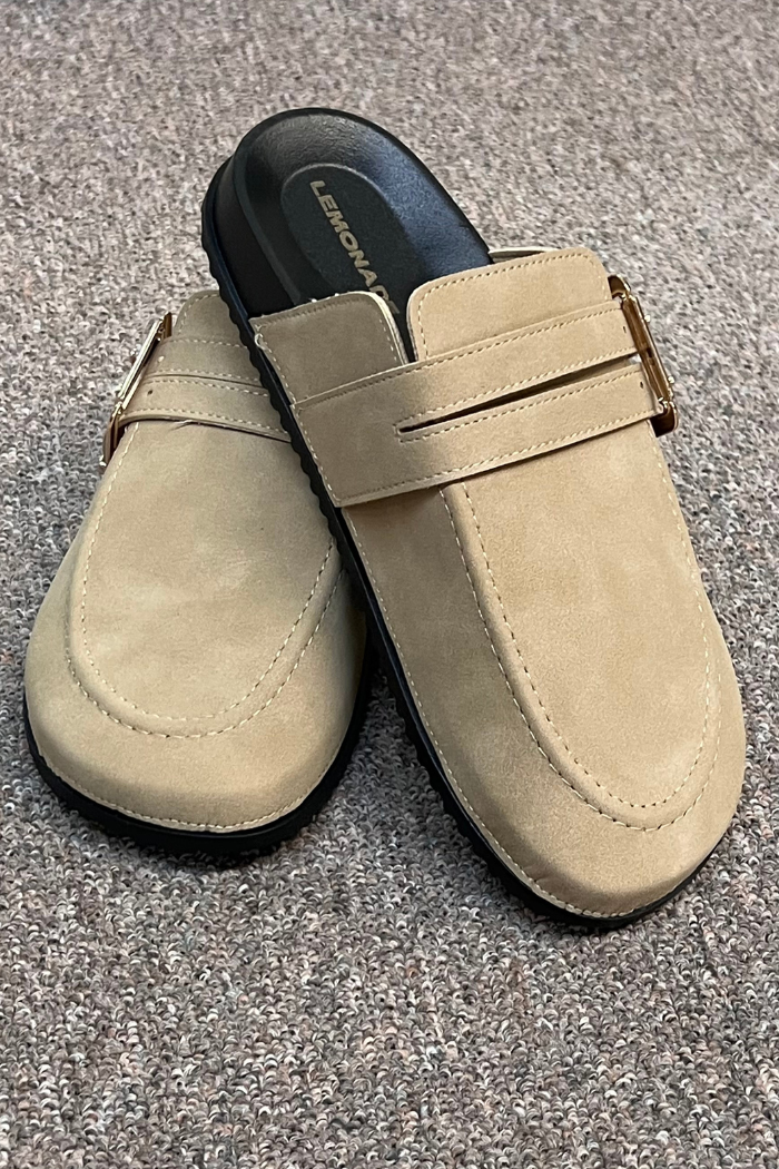A pair of Taupe Suede Clog Mules by Lemonade Shoes, featuring black soles and a gold buckle on the side, displayed on a textured carpeted floor.