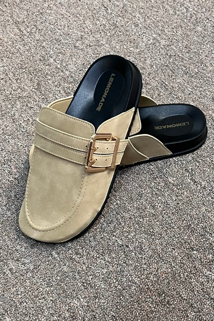 A pair of Taupe Suede Clog Mules with a gold buckle detail rests on a textured brown carpet. The LENMORAD branding is visible on the insole, reflecting the effortless style of Lemonade Shoes.