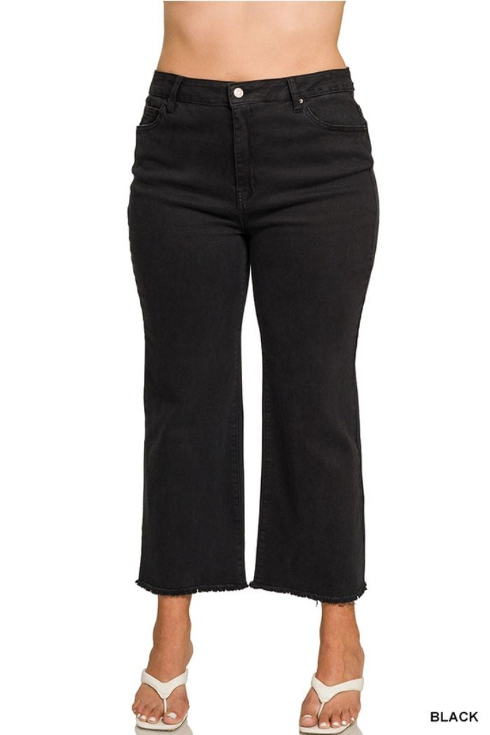 A person wearing Plus Acid Wash Pants, which are high-waisted, black cropped wide leg jeans with a raw hem and front pockets, styled with white heeled sandals. The pants feature a button and zipper closure.