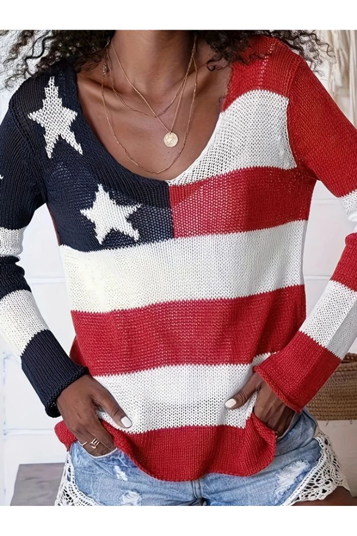 A person wears the American Flag Knit Sweater—featuring red and white stripes, navy with white stars—and light denim shorts with lace trim, hands tucked into the sweater’s pockets.
