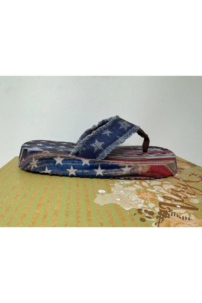 A Very G USA Flip Flop, featuring an Americana stars and stripes design in red, white, and blue, rests on a textured green and gold patterned surface.