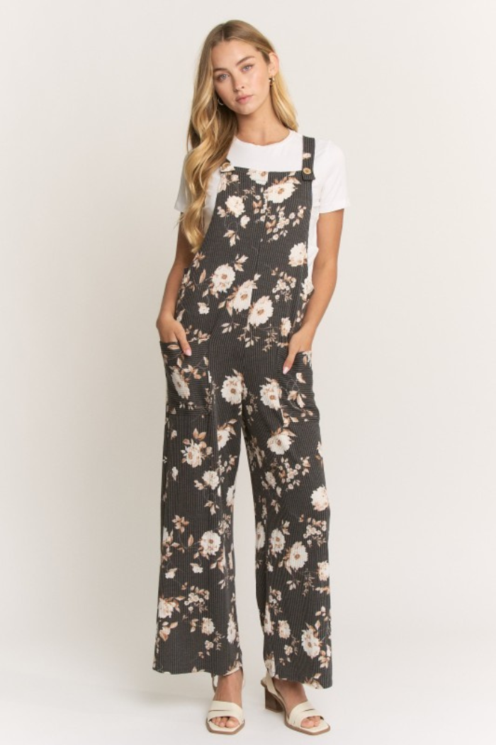 A woman with long blonde hair wears the Floral Ribbed Jumpsuit, featuring a stretchy, relaxed fit and wide legs, against a plain background. She pairs it with beige open-toe sandals and poses with her hands in the pockets.