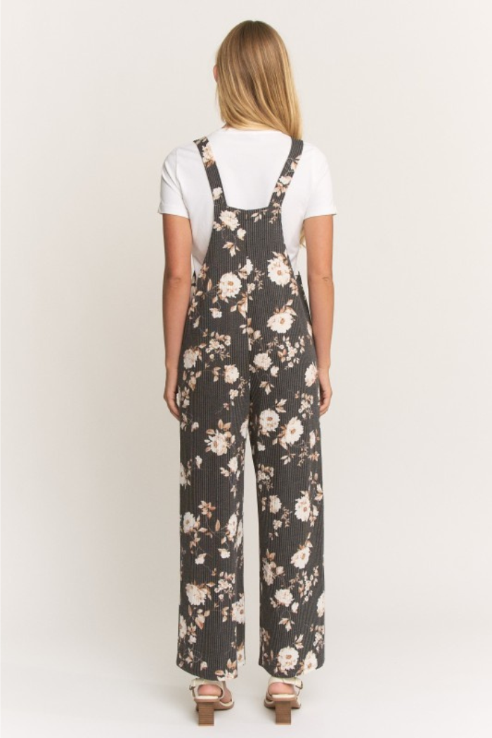 A woman with straight, light brown hair stands facing away in a black Floral Ribbed Jumpsuit with wide legs over a white short-sleeve shirt and light sandals, set against a plain white background.