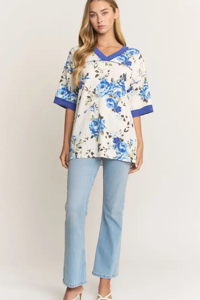 A woman with long, wavy blonde hair and a neutral expression wears the Floral Ribbed V-Neck Top featuring a blue floral print and trim, styled with light blue flared jeans and white heels.