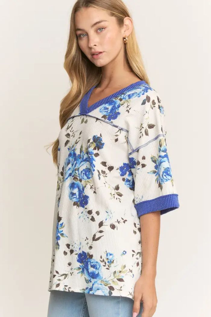A woman with long blonde hair wears the Floral Ribbed V-Neck Top, showcasing a blue floral print, blue trim on the sleeves and neckline, paired with light blue jeans against a plain background.