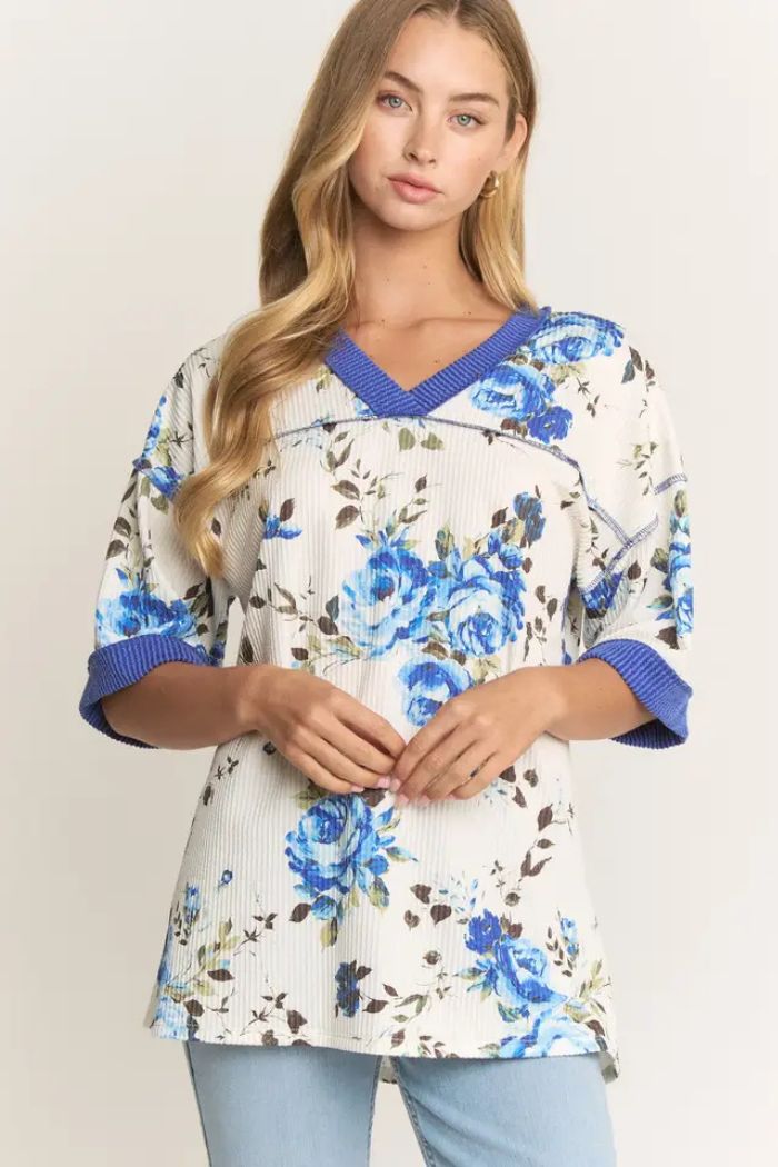 A woman with long, wavy blonde hair and a neutral expression showcases the Floral Ribbed V-Neck Top, featuring a blue floral print and solid blue cuffs, paired with light blue jeans for a relaxed look.