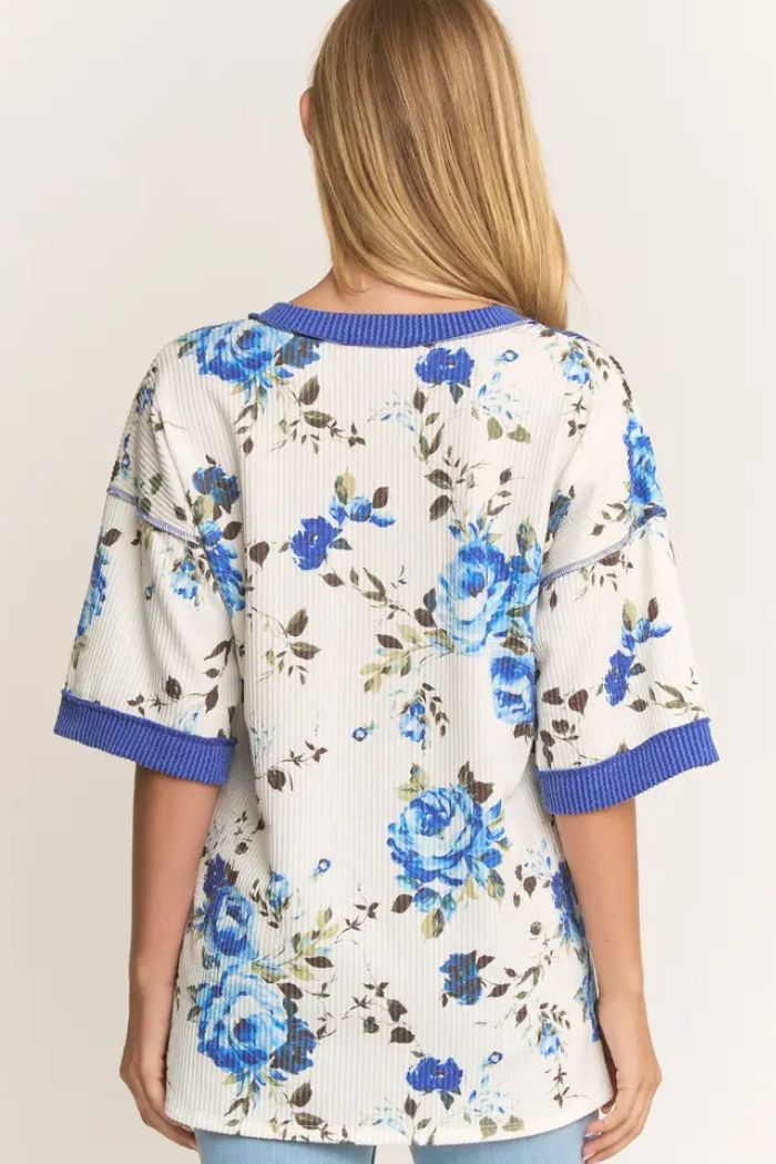 A woman with long blonde hair is seen from the back wearing the Floral Ribbed V-Neck Top in cream, featuring a blue floral print and wide blue trim on the sleeves and collar.