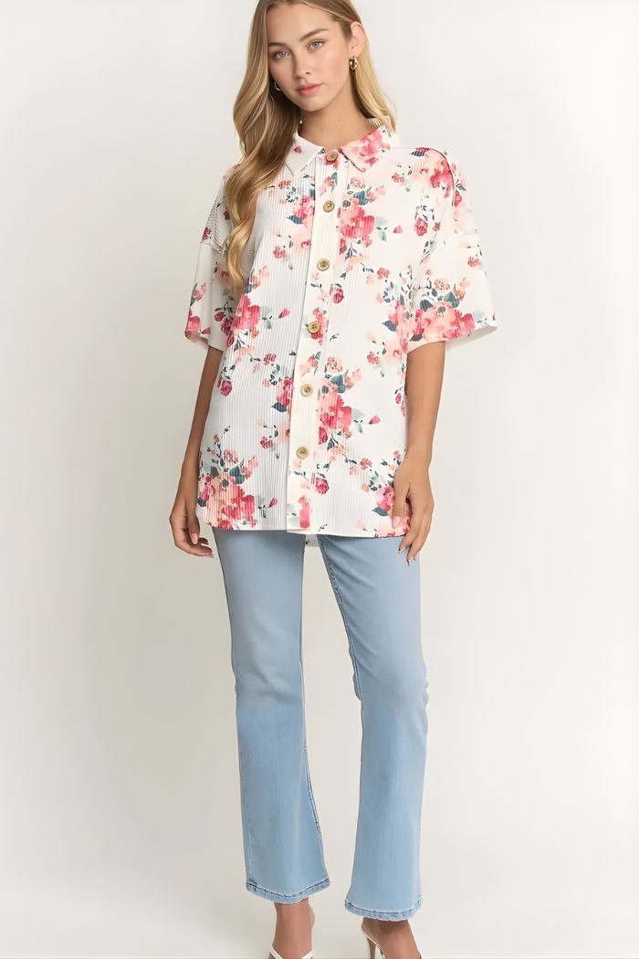 A woman stands against a plain light background wearing the Floral Short Sleeve Button Top, featuring a loose fit and watercolor floral print, paired with light blue straight-leg jeans and heels.