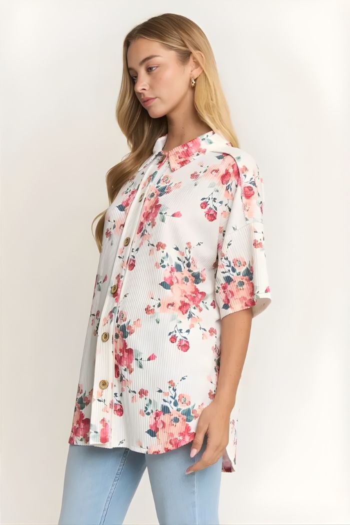A woman with long blonde hair wears the Floral Short Sleeve Button Top, featuring a pink watercolor floral print, and light blue jeans. She stands against a plain light background, gazing slightly downward and to the side.