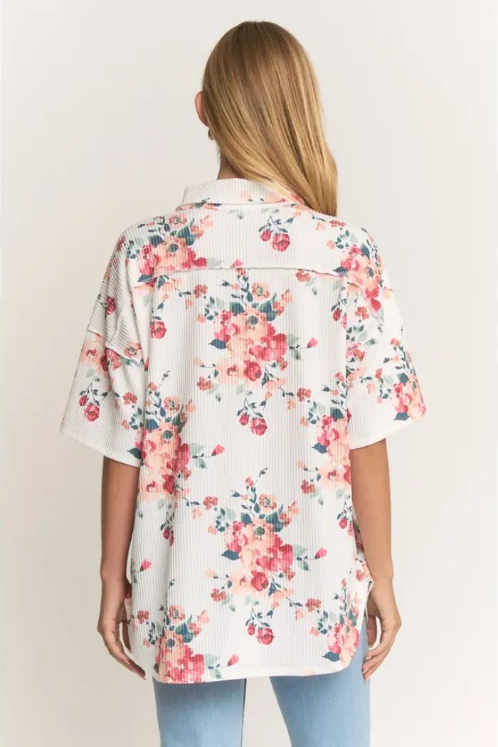 A woman with long, straight blonde hair is seen from behind, wearing the Floral Short Sleeve Button Top with a pink and orange watercolor floral print and light blue jeans.