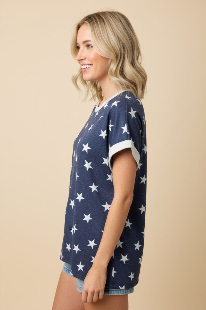 A woman with blonde, wavy hair models the Plus Star Terry Top, a navy blue star print top with white sleeve trim made from soft French terry knit, paired with denim shorts. She stands sideways, smiling against a beige background.